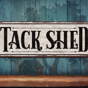 May include: A white metal sign with black lettering that reads "TACK SHED". The sign has a rustic, weathered look and is hanging on a blue wooden wall.
