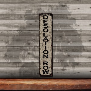 May include: A rustic metal sign with the words "DESOLATION ROW" in black lettering on a beige background. The sign is weathered and has a distressed look.