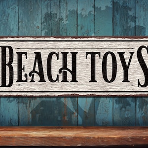 May include: A white sign with distressed edges and rusty brown paint. The sign reads "Beach Toys" in black lettering with a vintage font.