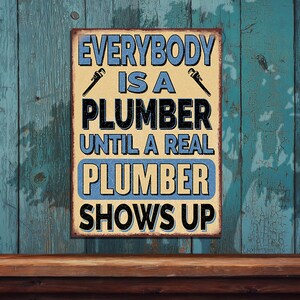 May include: A rectangular metal sign with the text "EVERYBODY IS A PLUMBER UNTIL A REAL PLUMBER SHOWS UP". The sign has a distressed look with a beige background, blue text, and wrench graphics. The sign is mounted on a weathered blue wooden surface.