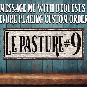 May include: A white sign with black lettering that reads "Le Pasture #9". The sign is made to look like old, weathered metal. The sign is hanging on a blue wooden wall.