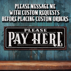 May include: A black and rusty metal sign with white text that reads "PLEASE PAY HERE". The sign is hanging on a blue wooden wall.