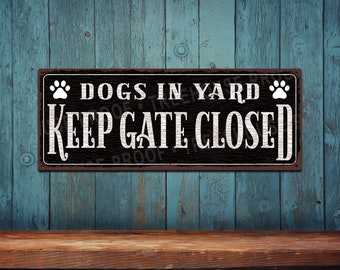 May include: A rectangular metal sign with a dark, weathered wood-grain background and a thin white border. The text reads "DOGS IN YARD" and "KEEP GATE CLOSED" in white, distressed lettering, flanked by two white paw print icons on the top corners.