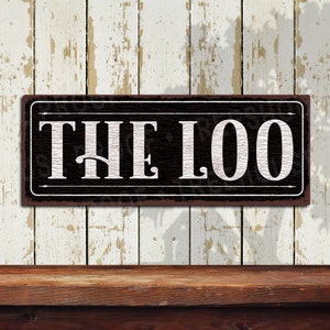 May include: A black and white metal sign with the text "THE LOO" in white letters on a black background. The sign is hanging on a white wooden wall.
