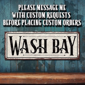 May include: A rectangular metal sign with the words "WASH BAY" in black, centered on a light wood-grain background. The sign has a rusty brown border and is mounted on a blue wooden wall. The text "PLEASE MESSAGE ME WITH CUSTOM REQUESTS BEFORE PLACING CUSTOM ORDERS" is above the sign.