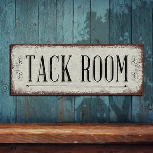 May include: A rectangular metal sign with the words "TACK ROOM" in black, centered on a white background. The sign has a distressed, vintage look with a rusty brown border and decorative flourishes. The sign is mounted on a weathered blue wooden wall.