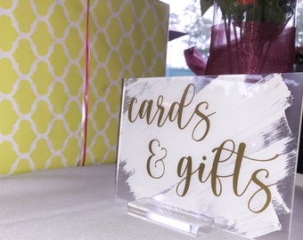 May include: A clear acrylic sign with the words "cards & gifts" in gold script, set on a clear stand. The background features a wrapped gift in yellow paper with a white geometric pattern and a pink ribbon.