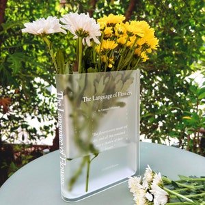 Acrylic book-shaped vase filled with vibrant flowers, placed on a modern bookshelf, perfect for home decor and gifting for events, birthdays, and housewarmings.