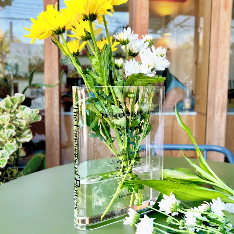 Acrylic book-shaped vase filled with vibrant flowers, placed on a modern bookshelf, perfect for home decor and gifting for events, birthdays, and housewarmings.