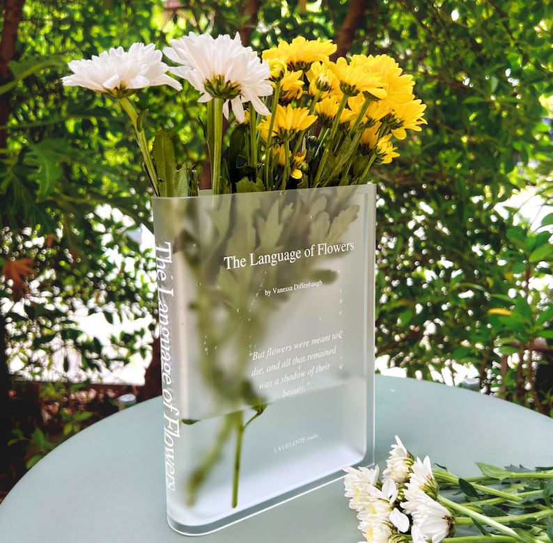 Acrylic book-shaped vase filled with vibrant flowers, placed on a modern bookshelf, perfect for home decor and gifting for events, birthdays, and housewarmings.