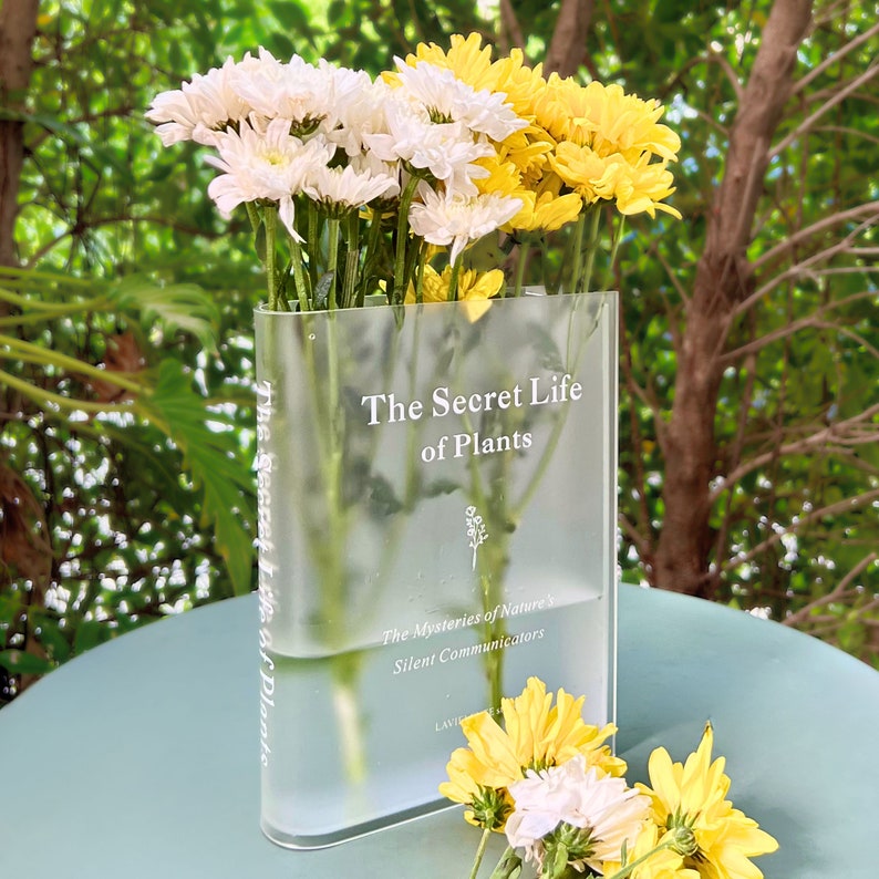 Acrylic book-shaped vase filled with vibrant flowers, placed on a modern bookshelf, perfect for home decor and gifting for events, birthdays, and housewarmings.