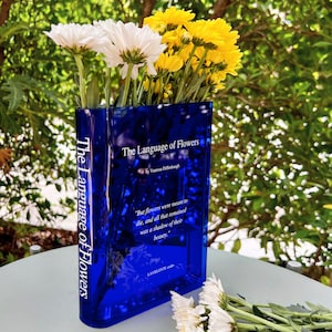 Acrylic book-shaped vase filled with vibrant flowers, placed on a modern bookshelf, perfect for home decor and gifting for events, birthdays, and housewarmings.