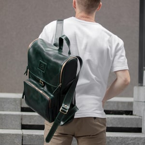 May include: A dark green leather backpack with a top handle, shoulder straps, and multiple compartments. The backpack has gold-colored zippers and accents. The person in the image is wearing a white t-shirt and khaki pants.