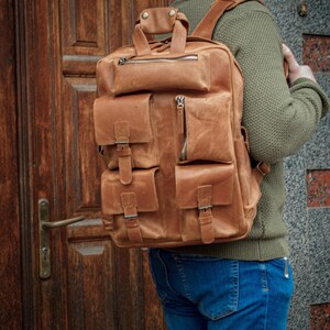 May include: A tan leather backpack featuring multiple exterior pockets with buckle closures, a top handle, and silver-toned metal zippers. The bag has a structured rectangular shape with visible stitching and adjustable shoulder straps for carrying.