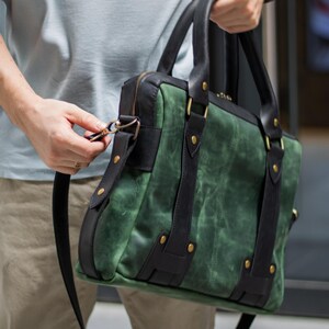 May include: Green and black leather briefcase with a shoulder strap. The bag features a textured green body with black leather accents, straps, and handles. Gold-colored rivets secure the straps. The bag is held by a person.