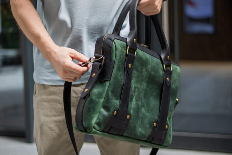 May include: Green and black leather briefcase with a shoulder strap. The bag features a textured green body with black leather accents, straps, and handles. Gold-colored rivets secure the straps. The bag is held by a person.