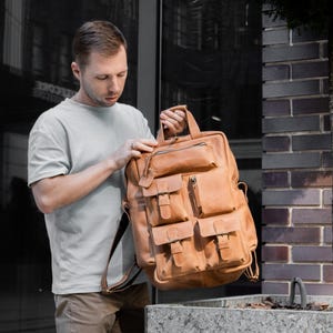 May include: A tan leather backpack with multiple front pockets and a top handle. The bag has adjustable shoulder straps and a zipper closure. The person in the image is holding the bag.
