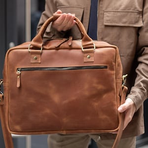 May include: A rectangular brown leather briefcase featuring two top handles, a detachable shoulder strap, and a horizontal zipped pocket on the front. The bag is constructed with brass-toned metal hardware, including rivets and zip pulls.