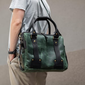 May include: Green and black leather satchel bag with a top handle and shoulder strap. The bag has black straps and gold-colored rivets. The bag is carried over the shoulder.
