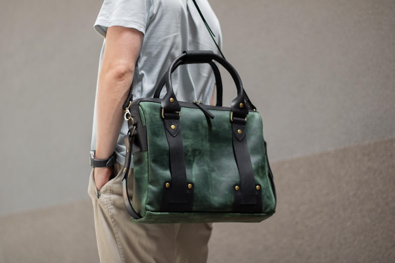 May include: Green and black leather satchel bag with a top handle and shoulder strap. The bag has black straps and gold-colored rivets. The bag is carried over the shoulder.