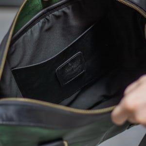 May include: Open view of a black leather bag with a green suede accent. The bag has a gold zipper and a black interior with a pocket. A small black leather tag with the word "Vintage" is visible inside the bag.