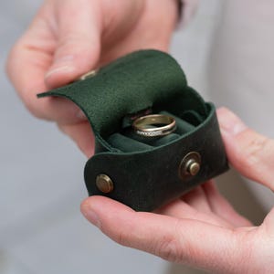 May include: A dark green leather ring box with a gold ring inside. The box is open, revealing a green velvet interior. The box is held in someone's hands and has a snap closure.