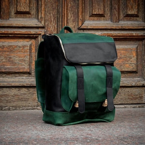 May include: A structured backpack crafted from dark green and black leather. The bag features a top flap, a large front pocket with two vertical black leather straps, and metallic buckles. The sides and base are green, while the main body is black.
