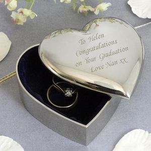 Op de afbeelding: Een zilveren hartvormige sieradendoos met een scharnierend deksel, open om een ring met een diamant te onthullen. Op het deksel staat de tekst "To Helen Congratulations on Your Graduation Love Nan XX". De binnenkant is gevoerd met donkerblauw fluweel.