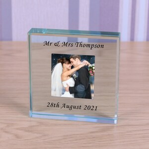 Puede incluir: Un marco de fotos de cristal transparente con una foto de boda de una pareja abrazándose. El marco tiene el texto "Mr & Mrs Thompson" y "28th August 2021" grabado.