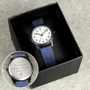 Può includere: Un orologio nero con quadrante bianco e cinturino in tela blu. L'orologio ha un retro argentato con un'iscrizione che recita "First Holy Communion Andrew Edwards 22nd July 2025" e una croce.