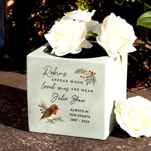 Può includere: Una fioriera commemorativa quadrata verde chiaro con rose bianche. La fioriera presenta il testo "Robins appear when loved ones are near Julie Shaw Always in our hearts 1967-2024" e un'illustrazione di un pettirosso.