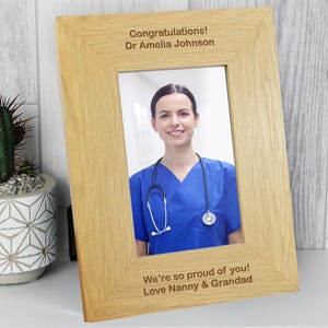 Può includere: Una cornice in legno di quercia chiara con il testo "Congratulations! Dr Amelia Johnson" in alto e "We're so proud of you! Love Nanny & Grandad" in basso. Contiene una foto di una donna sorridente in divisa blu e con uno stetoscopio.
