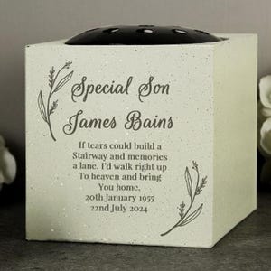 May include: A square, light-colored memorial flower vase with the text "Special Son James Bains." The vase includes a black floral insert on top and decorative floral accents. The inscription includes dates: January 20, 1955 - July 22, 2024.