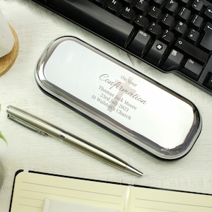 May include: Silver pen case with a white background and the text "On Your Confirmation" with a cross symbol. The text below reads "Thomas Jack Moore, 23rd July 2023, St Wulstan's Church".