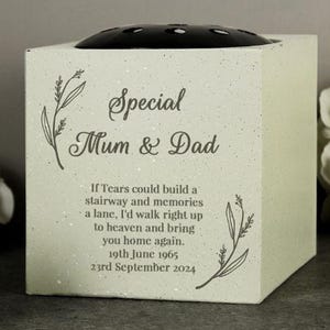 May include: A square, light-colored memorial vase with a black floral insert on top. The text "Special Mum & Dad" is displayed, along with a poem and dates. Floral line art accents the design.
