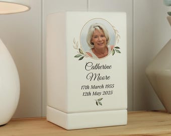 May include: A white memorial urn with a photo of a woman in a gold frame, surrounded by a green leaf design. The name "Catherine Moore" and dates "17th March 1955 - 12th May 2025" are printed below the photo.