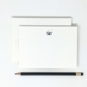 May include: A stack of white stationery cards with a small silver camera illustration on the top card. A black pencil is lying on a white surface below the cards.