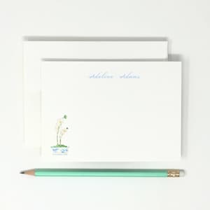 May include: A white stationery card with the name Adeline Adams printed in blue script at the top center and a small watercolor illustration of a white orchid in a blue and white patterned pot in the bottom left corner, paired with an envelope.