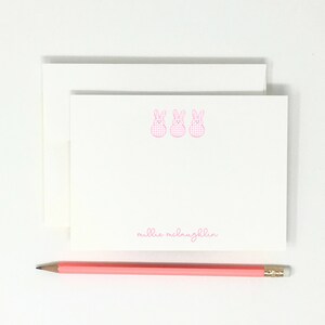 May include: White stationery set featuring three pink gingham bunny illustrations. The set includes a stack of notecards and envelopes, with the name "millie mclaughlin" printed in pink script. A pink pencil rests at the bottom.