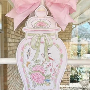 May include: A decorative, light pink door hanger shaped like a vase, adorned with floral and bird illustrations. A large pink bow is attached at the top. The vase design includes green accents and a bow. The overall theme is floral and decorative.
