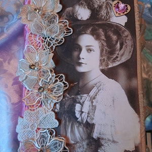 Puede incluir: Una foto vintage de una mujer con un gran sombrero y un vestido de encaje está pegada a una cubierta de diario de papel marrón. El diario está decorado con flores de encaje blanco y una cinta rosa. La foto tiene el texto "GG Ser.545/1" en la esquina inferior derecha.