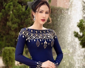May include: A woman wearing a dark blue dress with a beaded neckline and long sleeves. She is standing in front of a fountain with greenery in the background.