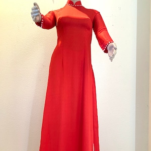 May include: A long, red dress with a high neckline and long sleeves. The dress has a white trim around the neckline and sleeves, and a white slit up the front.