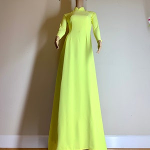Puede incluir: Un vestido largo de manga larga de color amarillo brillante con cuello mandarín. El vestido es de largo hasta el suelo y tiene un diseño simple y elegante.