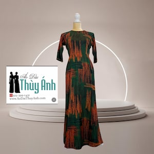 Op de afbeelding: Een lange, groene jurk met een patroon van oranje penseelstreken wordt getoond op een mannequin. De jurk heeft driekwart mouwen en een ronde halslijn. Een logo met tekst is zichtbaar aan de linkerkant van de afbeelding.
