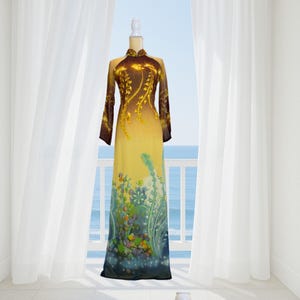 Op de afbeelding: Een lange, gele jurk met een bloemenontwerp en lange mouwen. De jurk heeft een hoge kraag en wordt op een mannequin getoond. De achtergrond toont een balkon met uitzicht op de oceaan, met witte gordijnen aan weerszijden.