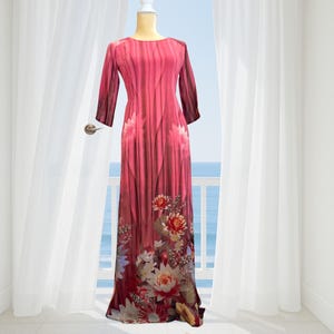 Op de afbeelding: Een lange jurk met lange mouwen in rood- en bourgondietinten, versierd met een bloemenpatroon. De jurk heeft een ronde hals en wordt gepresenteerd op een mannequin. De achtergrond toont een balkon met uitzicht op de zee.