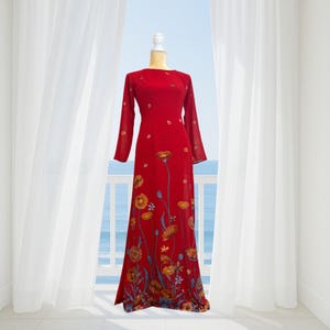 Op de afbeelding: Een lange, rode jurk met lange mouwen en een bloemenpatroon. De jurk heeft een ronde halslijn en wordt op een mannequin gepresenteerd. Het bloemmotief bevat oranje en gele bloemen met blauwe accenten. De jurk is vloerlang.