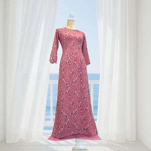 Op de afbeelding: Een lange, stoffige roze jurk met een bloemenpatroon. De jurk heeft lange mouwen en een ronde halslijn. Het wordt gepresenteerd op een paspop voor een raam met uitzicht op de zee.