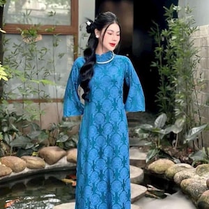 May include: A long, blue dress with a mandarin collar and flared sleeves. The dress has a repeating circular pattern. The person wearing the dress has long, dark hair. The background includes greenery and a water feature.